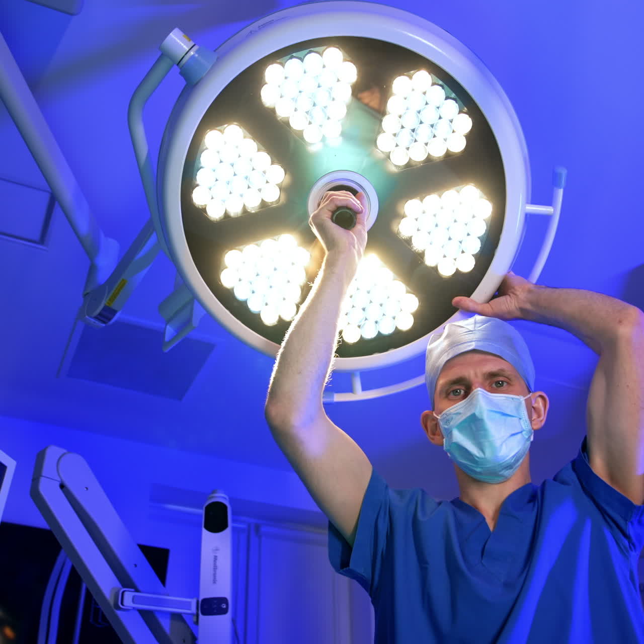 Médico asistente enciende la gran lámpara redonda en el techo de la sala de cirugía. La lámpara encendida ciega la cámara. Vista en ángulo bajo.