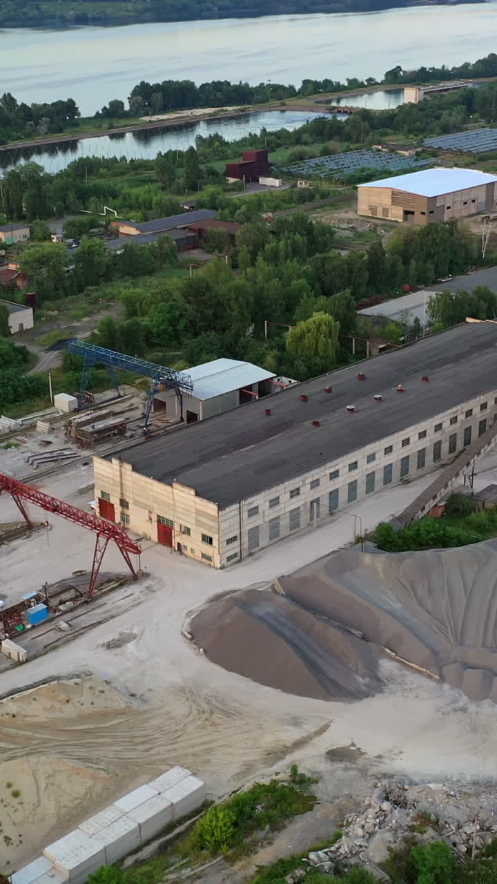 Construction site from bird's eye near the river. Buildings of industrial factory on the bank of the river. Aerial view. Vertical video