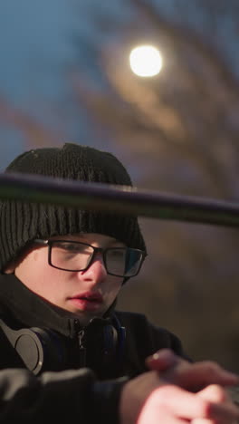 close-up of a young boy wearing glasses and a black jacket resting on an iron bar with hands clasped together, with blurred lights and bare trees in the background