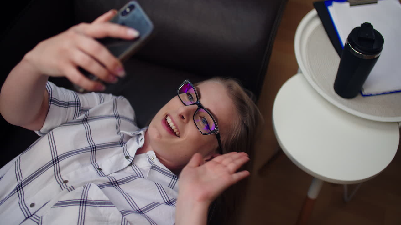 atractiva mujer de negocios que tiene una videollamada en el teléfono móvil mientras está acostada en el sofá en la oficina 1