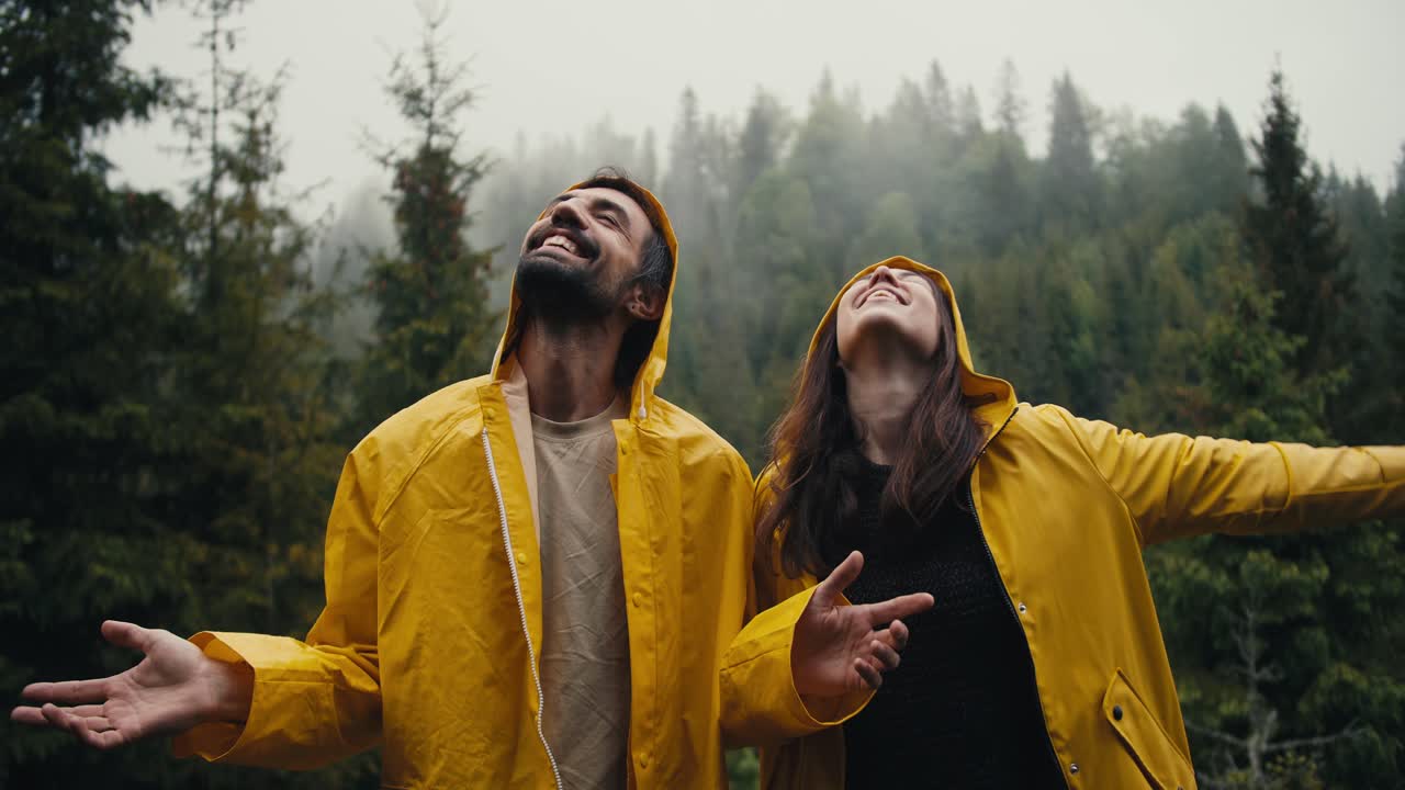Happy couple: a guy and a girl in yellow jackets look at the sky, smile and wait for the coming rain against the backdrop of a mountain coniferous forest