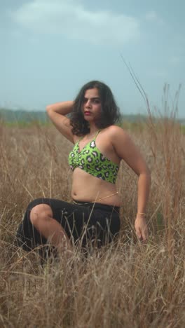 a woman kneels in tall dry grass wearing a green leopard-print top, striking a confident pose outdoors