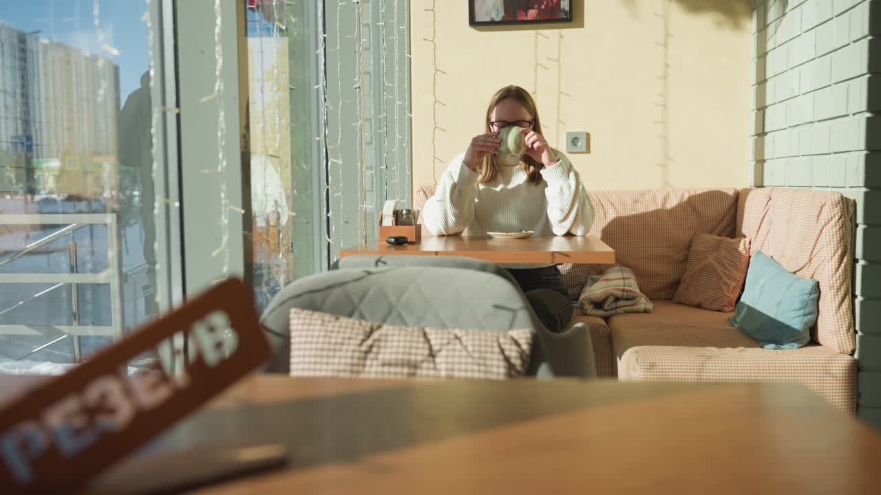 Front view of young woman in glasses sipping morning tea inside cozy cafe, seated by window with soft pillows, with reflections of people walking and snowy urban street scene in background