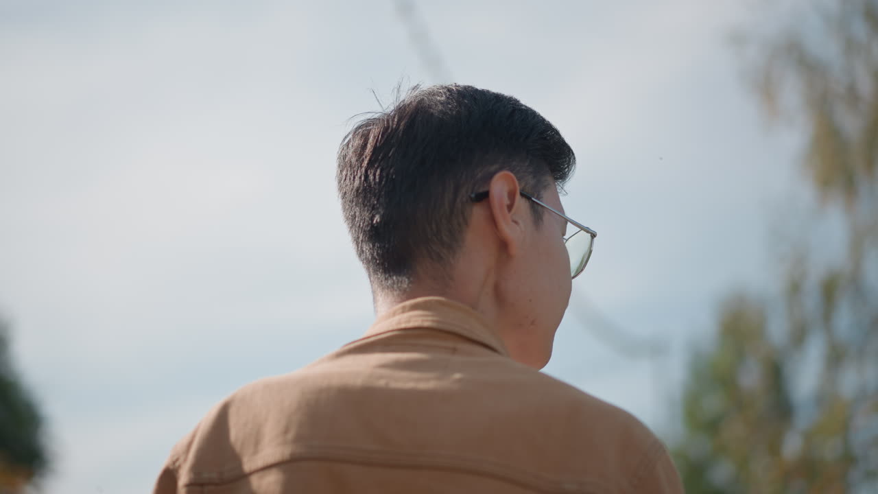 uomo asiatico di spalle che vaga per la strada, vista posteriore di un giovane uomo asiatico che cammina sotto un cielo blu con fili e alberi, giacca marrone e occhiali creano una silhouette silenziosa, pali suburbani e fili luminosi