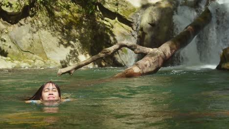 young woman swimming at the base of a waterfall in the caribbean in a blue bikini