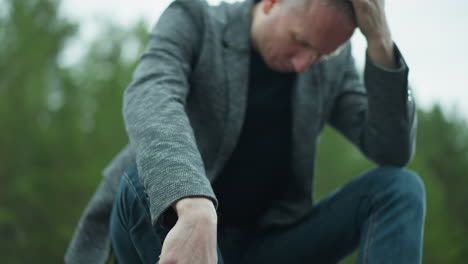 a downward view of a man in a gray jacket and blue jeans, wearing canvas shoes, handling a handgun while sitting beside a railway track, his head is bowed, and one hand is on his head