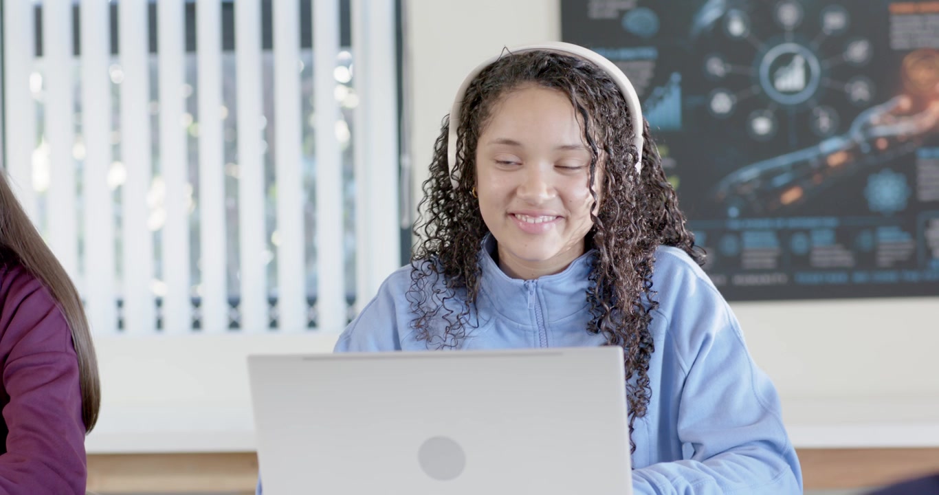 Diverse teens in class working on laptops, message on her laptop making smile while wearing headset