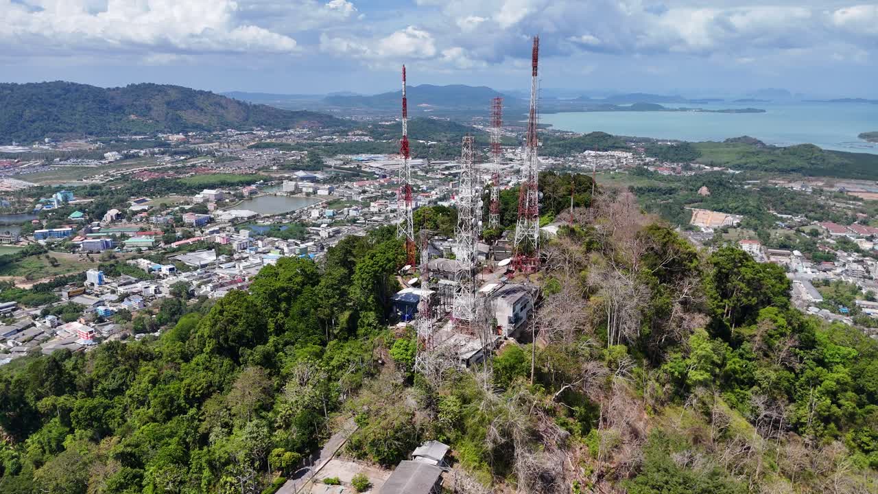 드론 영상은 태국 푸켓의 원이 언덕 꼭대기에 있는 통신 탑을 포착합니다. 은 하늘 아래 울창한 초록색과 도시 풍경으로 둘러싸여 있습니다.