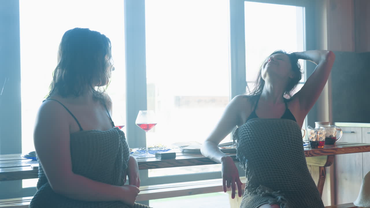young woman wrapped in towel setting cup on breakfast table in rustic kitchen while looking toward friend in soft natural morning light from large window