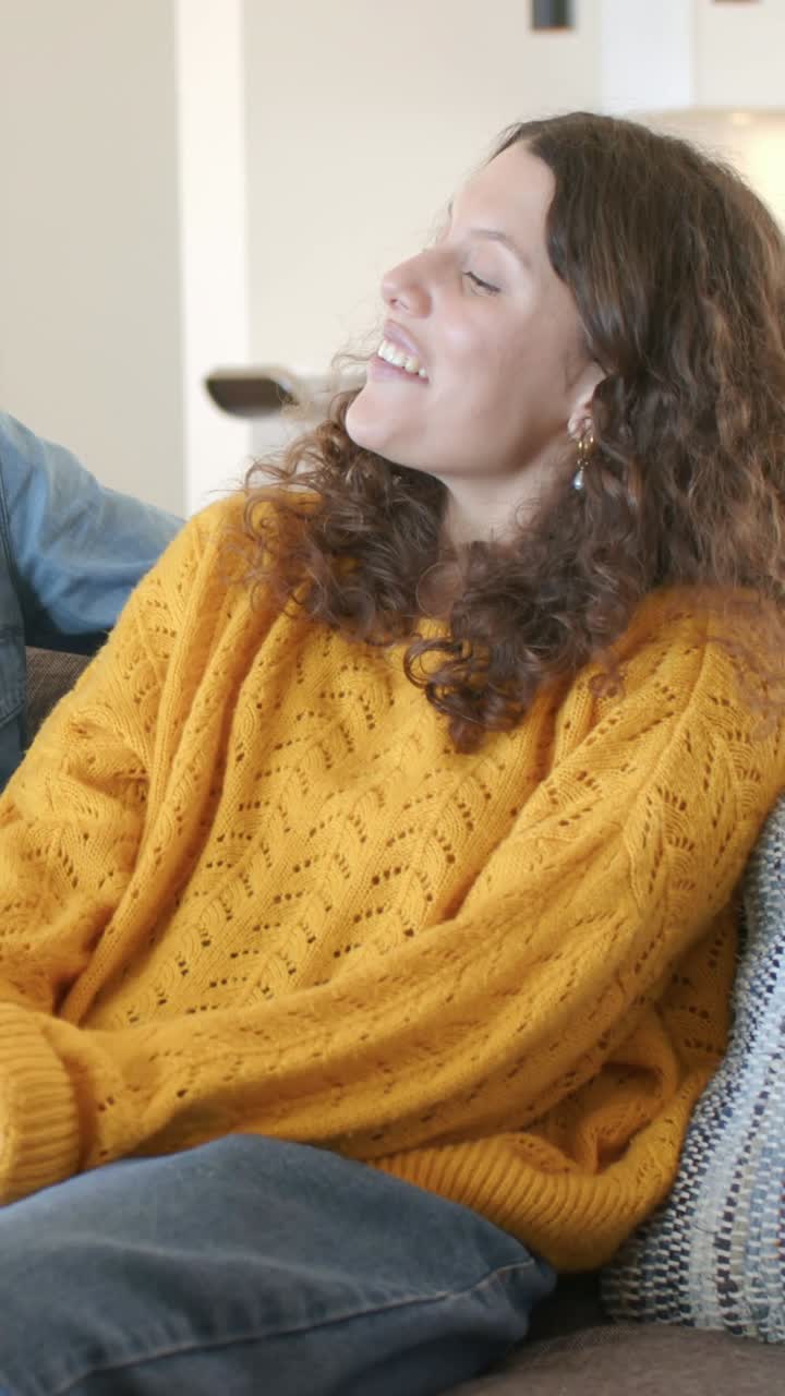 Vídeo vertical: Mulher rindo, com suéter mostarda e jeans, em casa, ouvindo um(a) amigo(a) contar uma piada e depois falando.