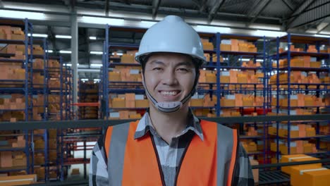 primer plano de un ingeniero asiático con casco de seguridad sonriendo a la cámara mientras está de pie en el almacén con estantes llenos de mercancías de entrega