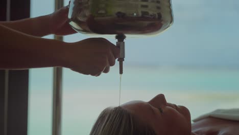 masajista comenzando el flujo de aceite caliente del cuenco en la frente de la mujer, shirodhara