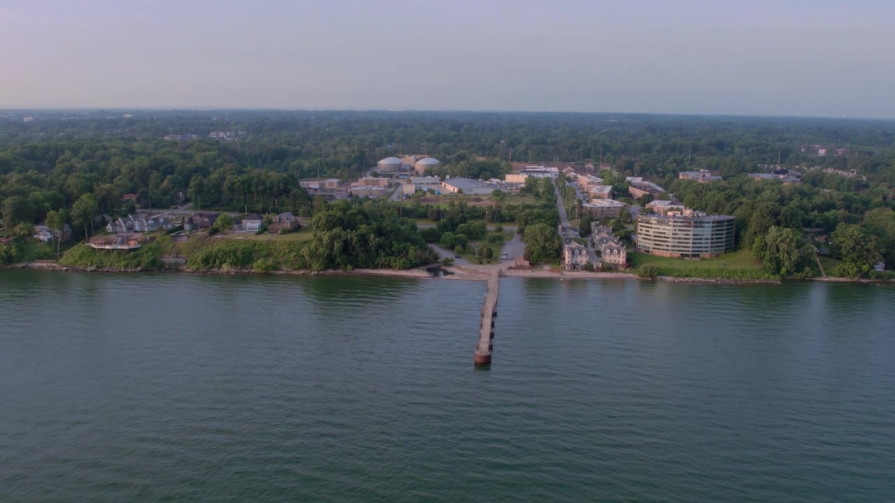 Drone sulla spiaggia del Lago Erie sopra l'acqua rivolto a sud, carrellata inversa e panoramica verso l'alto, mostrando alberi verdi, quartieri residenziali ed edifici per uffici