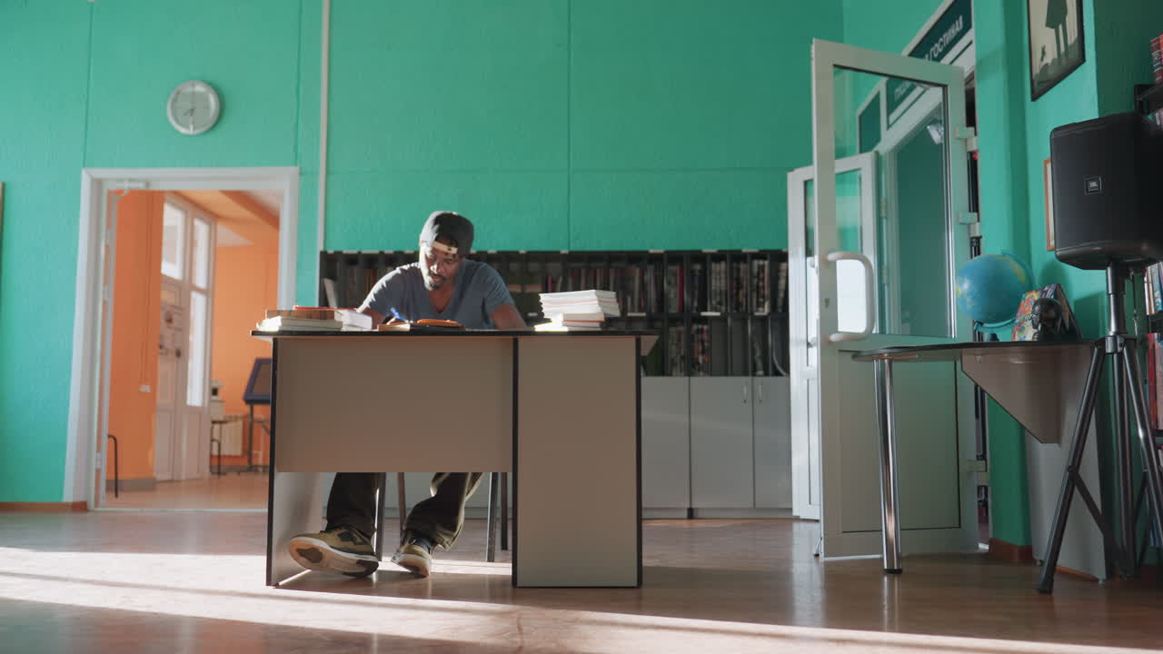 Academician sits at desk in brightly lit library, writing with pen while flipping to new page, surrounded by books and shelves, focused on work as sunlight streams across floor