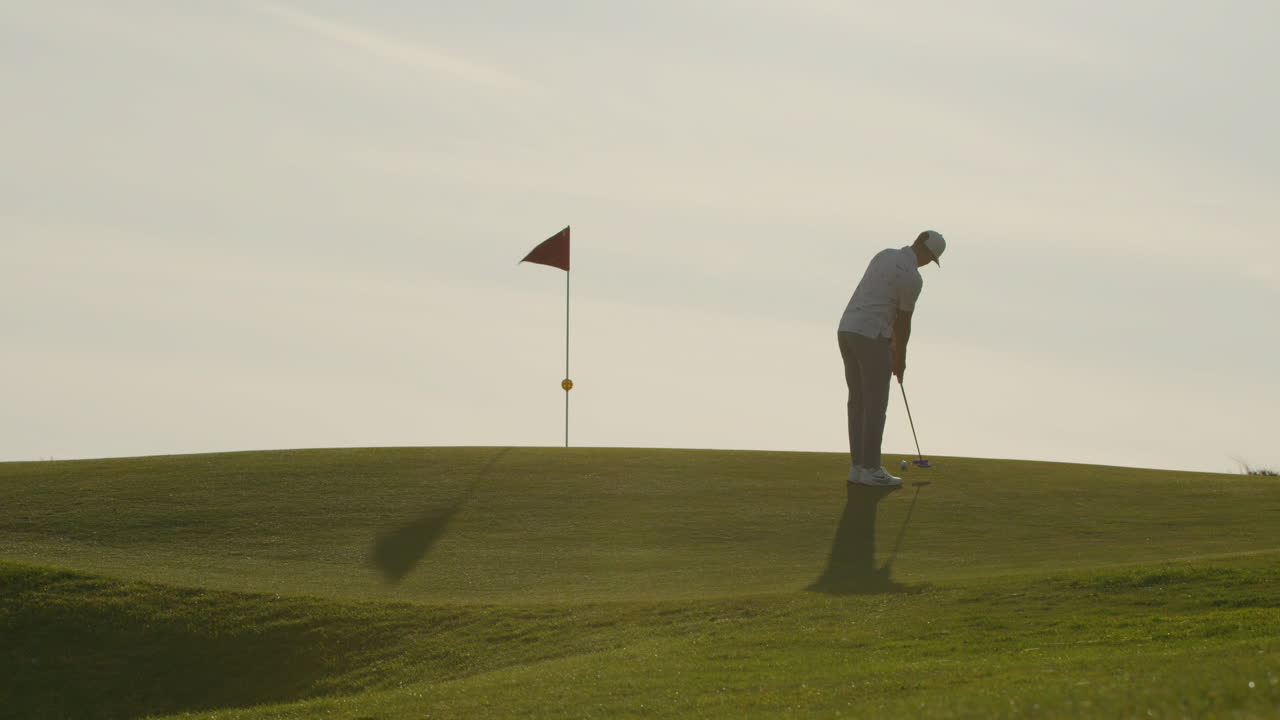Golfista uomo che imbuca una pallina da golf su un green al tramonto.