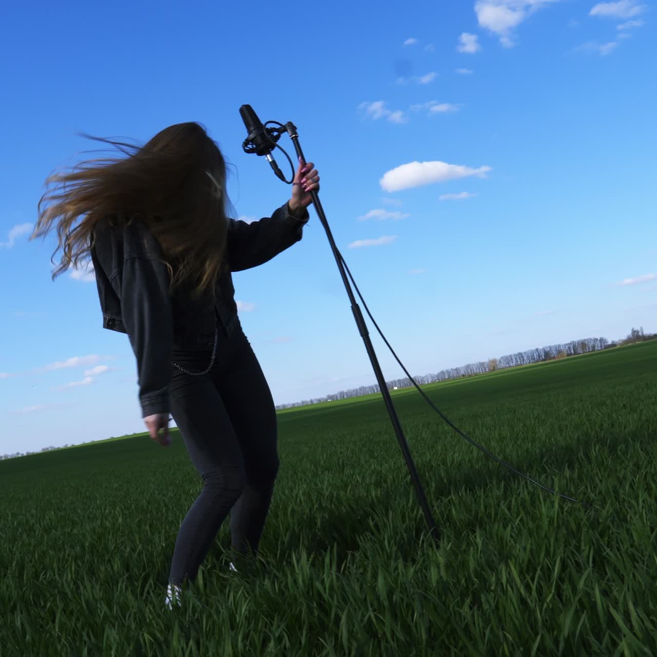 Cheerful happy young lady dancing emotionally near the microphone stand in the green field. Female having fun while making video for music clip