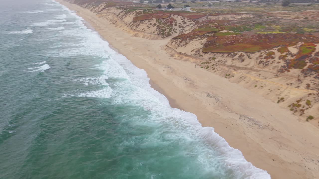 Drone starts above ocean facing dunes, rotates and flies parallel to the beach, tracking coastline