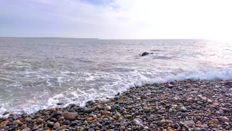 Zeitlupenwellen-Brechen-An-Einem-Farbenfrohen-Steinstrand-Mit-Einer-Landzunge-Im-Hintergrund