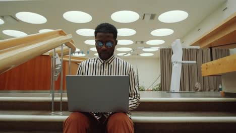 Afroamerikanischer-Mann-Sitzt-Auf-Der-Treppe-In-Der-Bibliothek-Und-Benutzt-Einen-Laptop