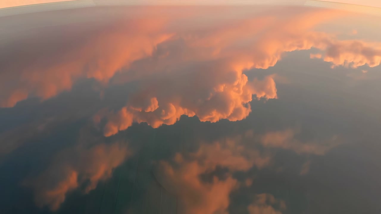 Soft sunset light illuminates dramatic orange cloud reflections across a smooth dark surface, with a curved metallic edge at the top and a hazy atmospheric