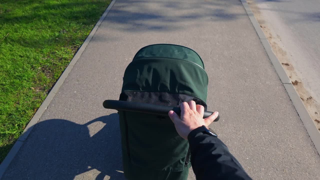 A person pushing a green baby stroller along a sunny sidewalk in a park. The scene captures a peaceful walk with a view of the stroller and shadow on the pavement.