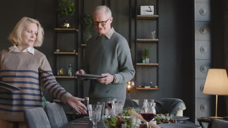feliz pareja de ancianos poniendo platos en la mesa del comedor, mientras que en el fondo su familia se sienta en la sala de estar y habla juntos