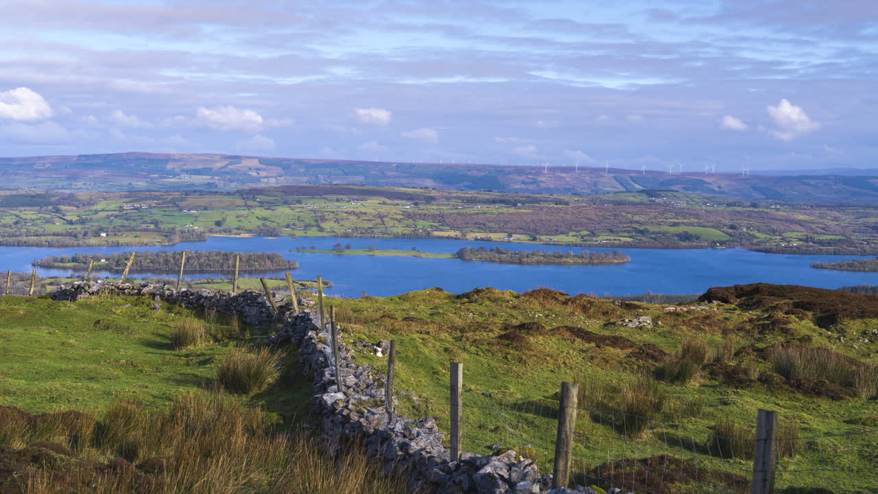 Timelapse di un paesaggio rurale con muro in pietra e pilastri in legno in un campo erboso, colline e lago in lontananza durante una giornata nuvolosa, visto da Carrowkeel, nella contea di Sligo, in Irlanda