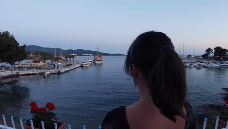 steady camera movement from the back of a young girl wearing a dress and then looking at the camera while standing on a marine in a small greek town on sunset