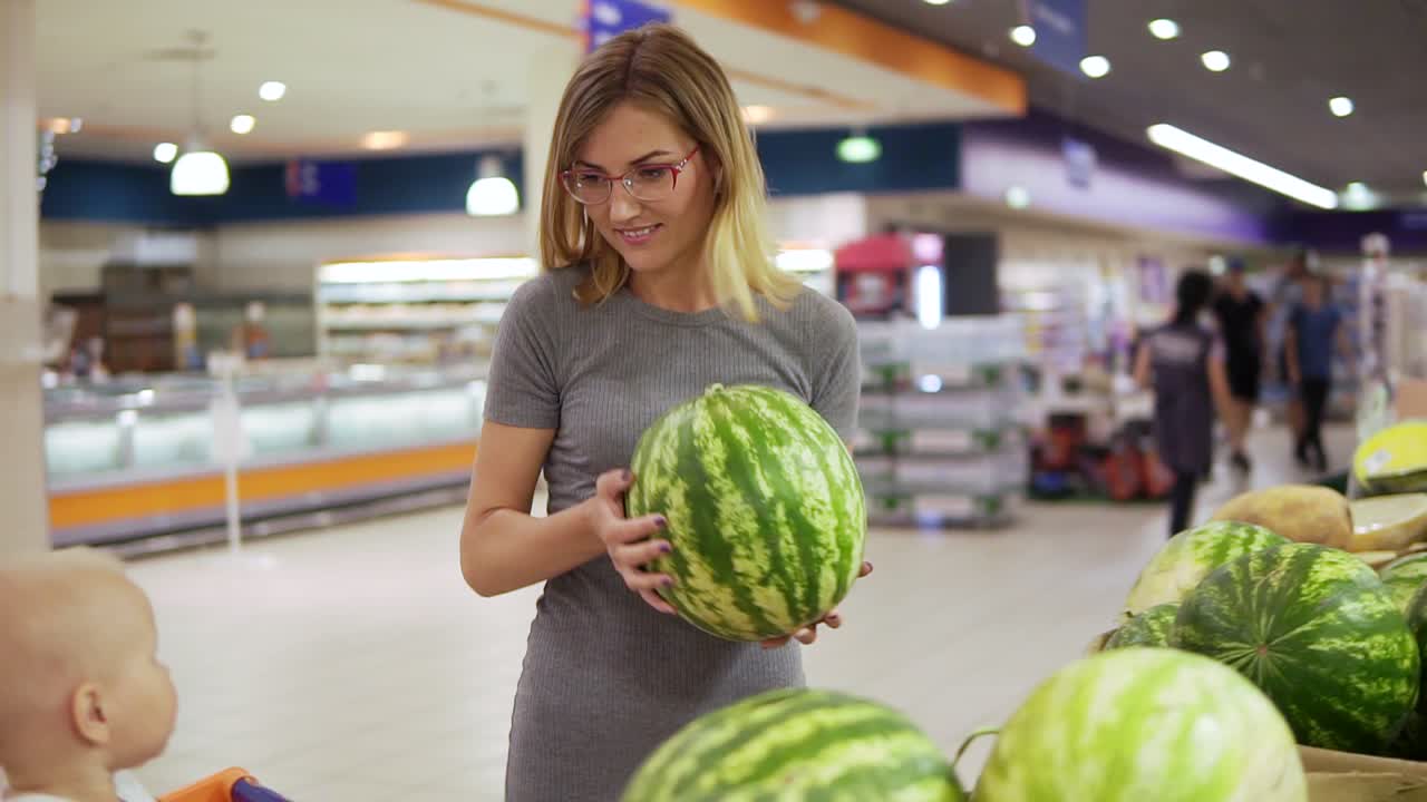 Cute little child is helping her mother to choose a watermelon in the supermarket. Young mother is showing her baby a water