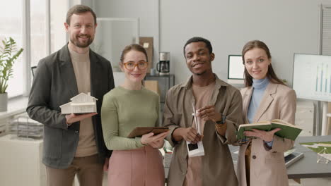 multiethnic group posing at camera holding wind turbine, house model, notebook and tablet