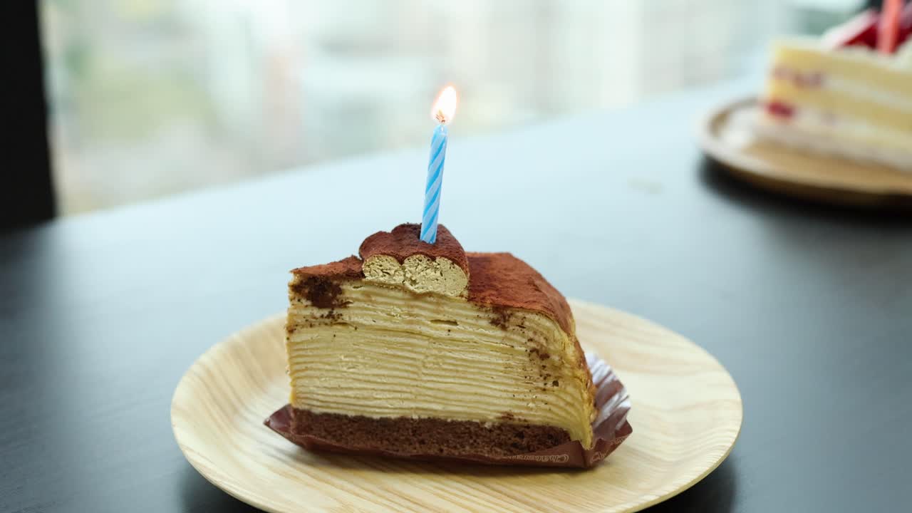 Hand lights blue candle on tiramisu crepe cake slice in natural daylight, close-up view
