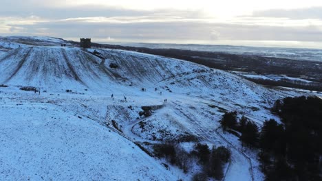 Snowy-Rivington-Pike-tower-Winter-hill-aerial-view-people-sledding-downhill-at-sunrise-forward-moving