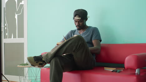 young man in cap prepares to sit on red couch in library lounge, sets books in hands for quiet study session, looking down at seat, relaxed mood