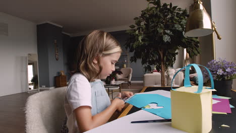 Side-View-Of-Girl-Cutting-Heart-Shaped-Cardboard-On-Desk-In-Living-Room