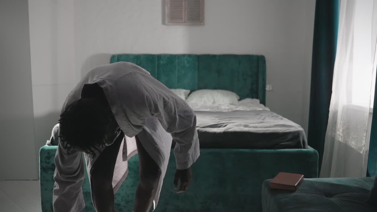 Man in bathrobe holding cup of tea while walking past bed in modern bedroom. Green velvet bed with pillows, cozy setting with natural light streaming through window