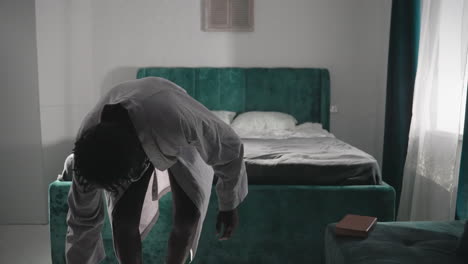 man in bathrobe holding cup of tea while walking past bed in modern bedroom. green velvet bed with pillows, cozy setting with natural light streaming through window