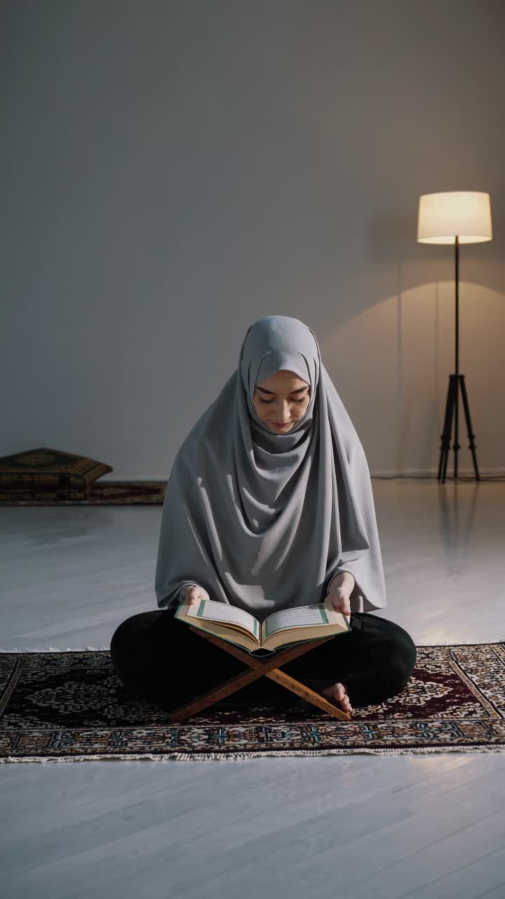 Sitting on a prayer rug, a young Muslim woman in a hijab reads the Quran, bathed in soft light from a lamp, creating a serene atmosphere of devotion