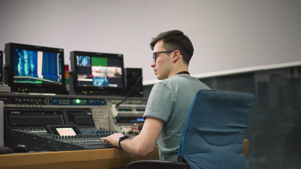 Broadcast operator working in control room, managing audio and video mixing equipment while monitoring live broadcast on computer screens and handling various broadcasting tasks in television studio