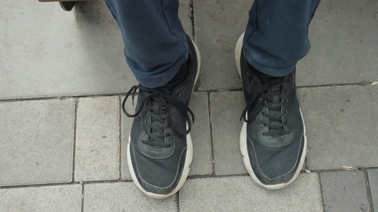 Dengeli duruşu ve hazır adımı yakalayan kaldırım üzerindeki spor ayakkabıların yukarıdan görünümü, denim pantolon, bağlı bağcıklar, açılı eşleştirilmiş ayaklar, sabah işe gidip gelme havası, belirgin karo deseni, yakın çekim ayakkabı detayı