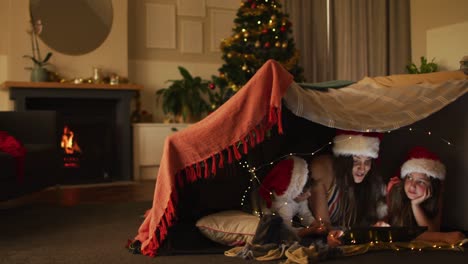 Happy-caucasian-mother-and-siblings-wearing-santa-hats-lying-in-makeshift-tent,-using-tablet