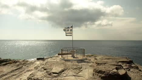 wide shot of a greek flag waving in slow motion with the sea in the background, on a rocky shore