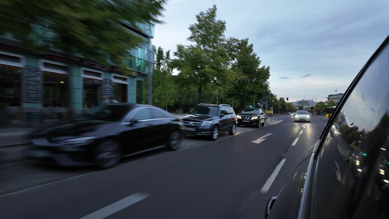View from a moving vehicle along a Berlin street. A row of parked cars lines the street. Dynamic, urban atmosphere.