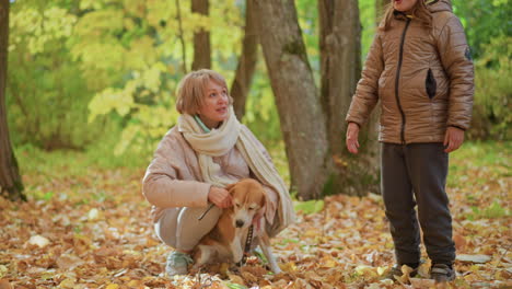 little girl running through golden leaf carpet in autumn forest holding dog leash out to kneeling mother wearing coat and jeans, eager beagle sitting among trees in playful handover moment