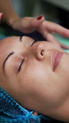 caucasian woman wearing protective cap lies with eyes closed on the couch at salon. cosmetologist applying some gel from a little glass cup on client's face. close up. vertical video