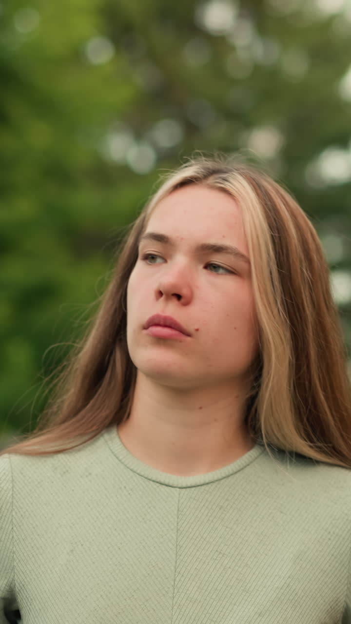 mujer joven caminando por el camino al lado de la valla de hierro, con una expresión pensativa y de mal humor, mirando ligeramente aburrido, rodeado de exuberante vegetación y estructuras de patio de recreo en el fondo
