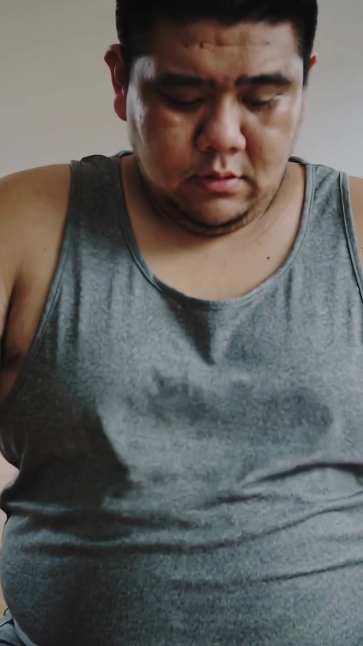 Video vertical: Hombre sentado bajando la mirada en un asiento de una habitación, vistiendo una camiseta sin mangas gris.