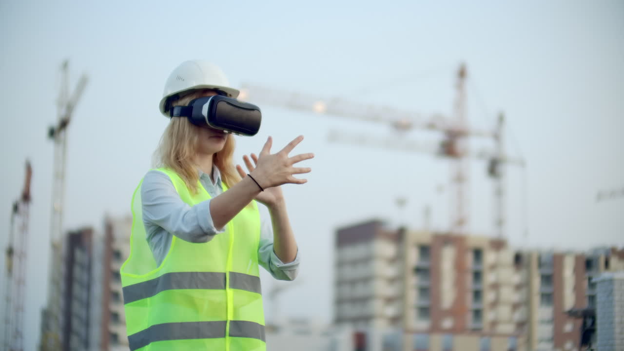 mujer diseñadora en un sitio de construcción en casco duro y chaleco en las gafas de realidad virtual para mover sus manos imitando la interfaz en el fondo de las grúas al atardecer.