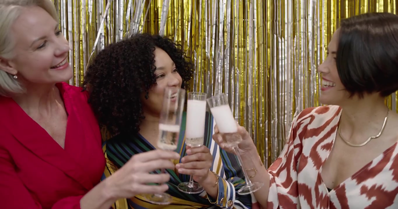 Three women by gold fringe, right woman pouring green bottle, center woman reacting