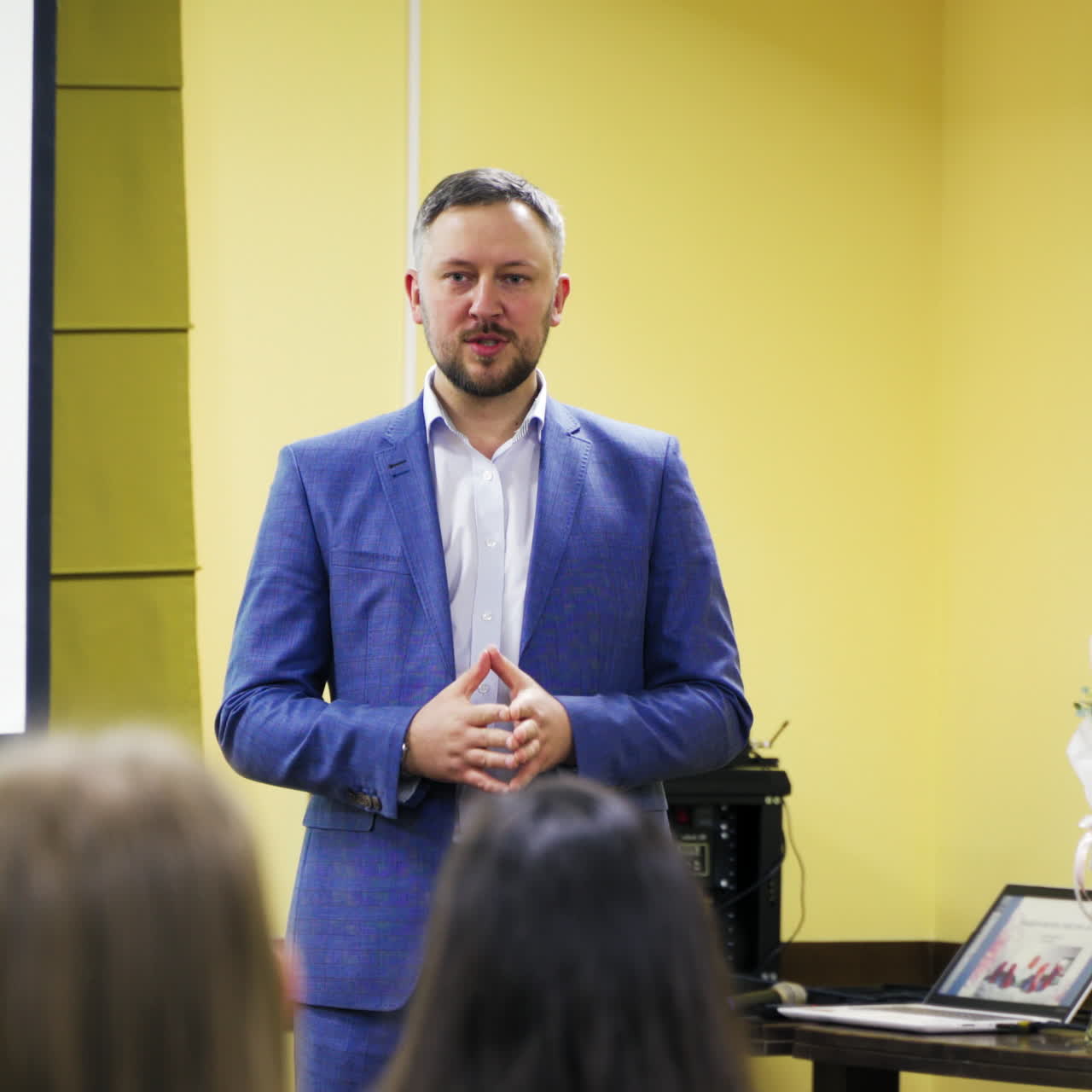 Successful leader is conducting a lecture while standing near table with flowers and a laptop in hall. Businessman in blue suit speaks at the business forum for public people.