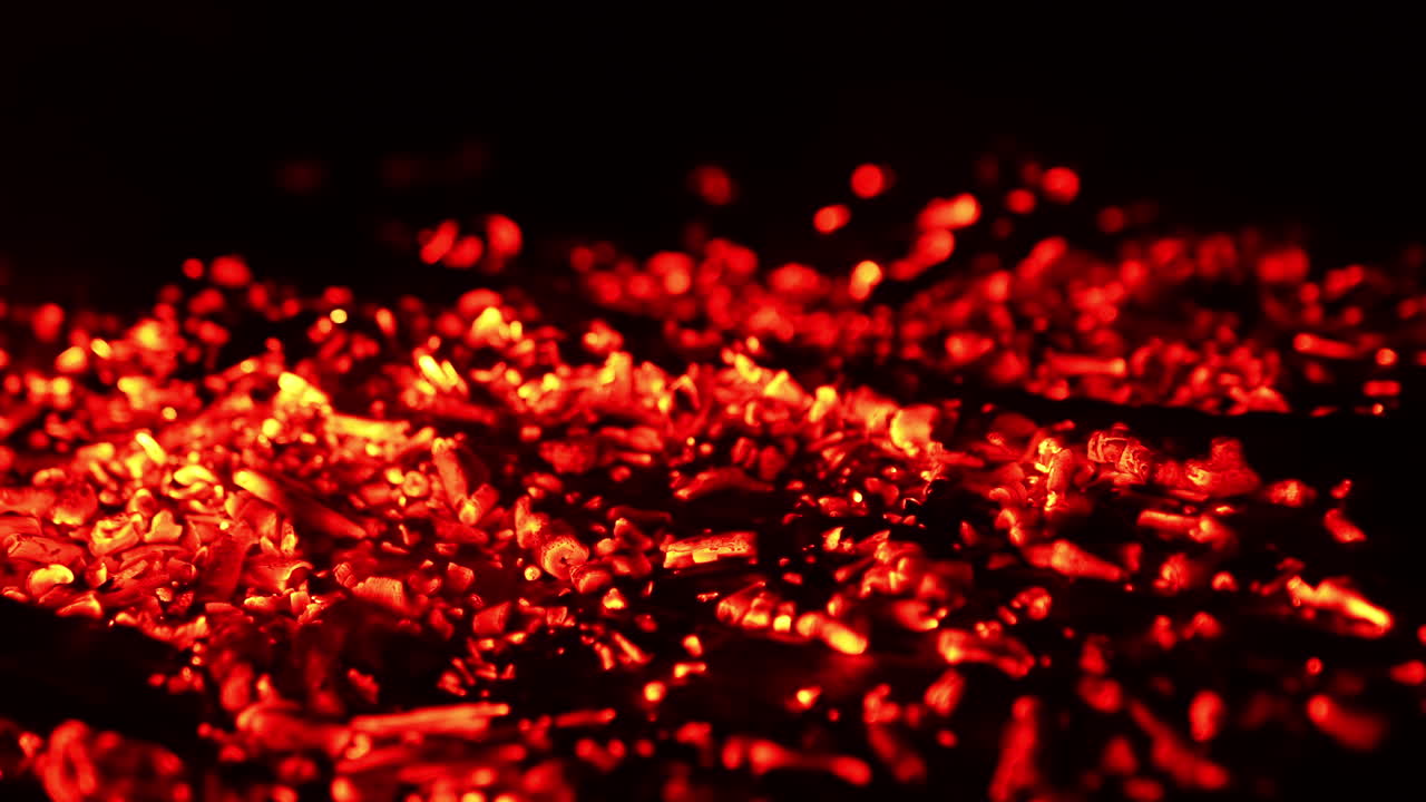 Smoldering red embers scattered on the ground. Smoke goes up and charcoal flashing from the wind. Close up.